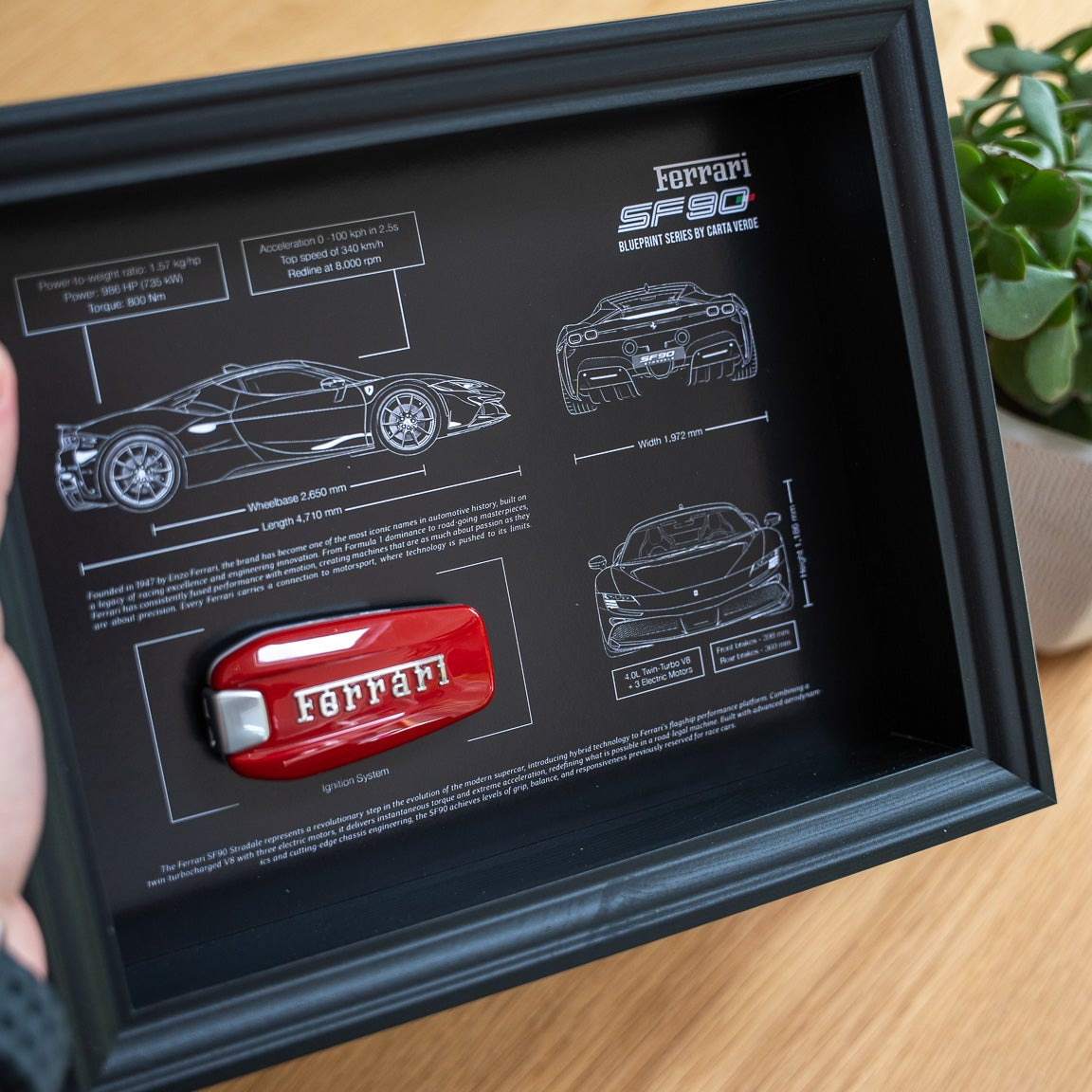 Ferrari car key in a black frame with technical drawings on a wooden surface.